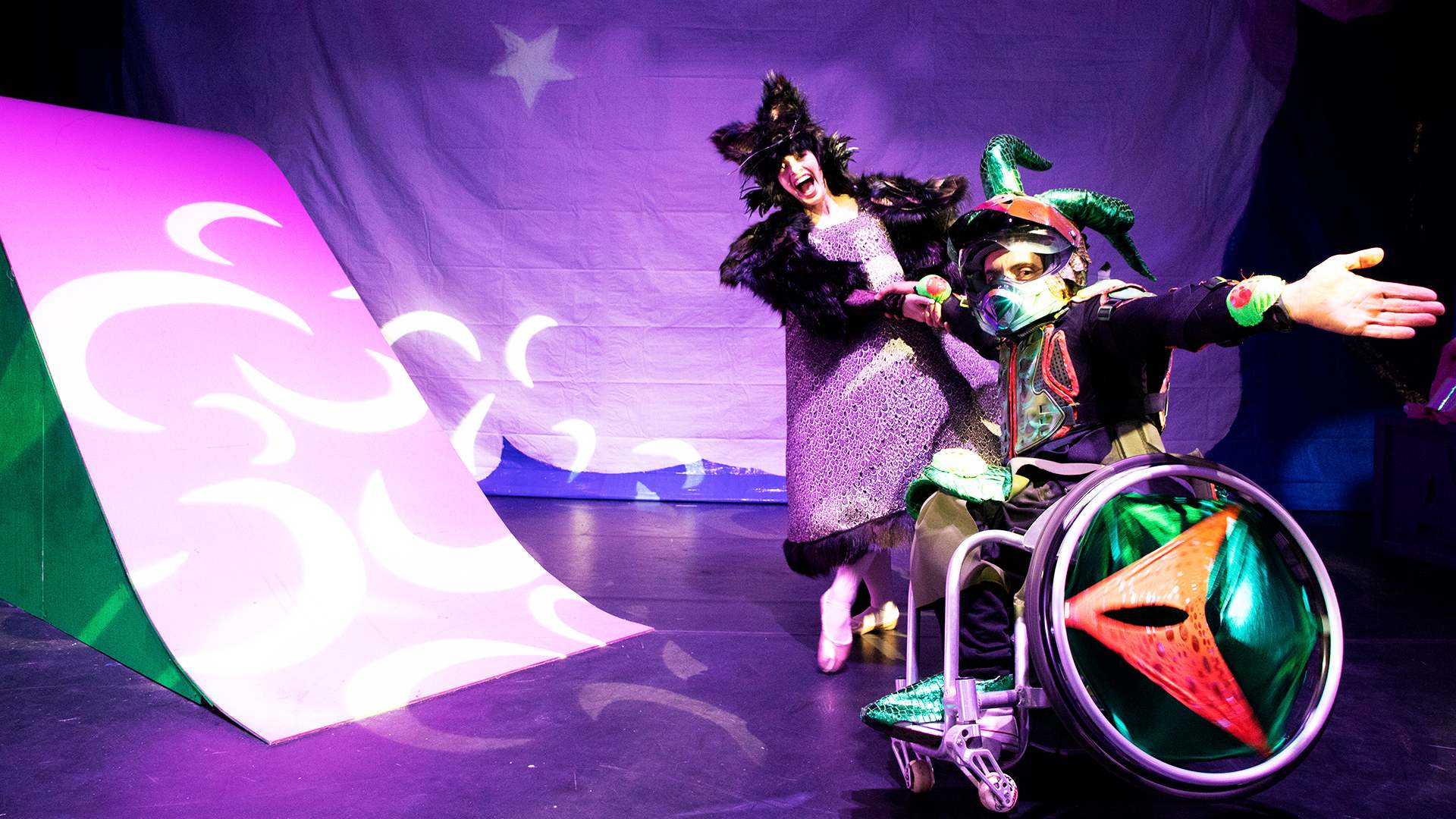 Performer in a wheelchair and performer standing both wearing costumes for She Conjured the Clouds