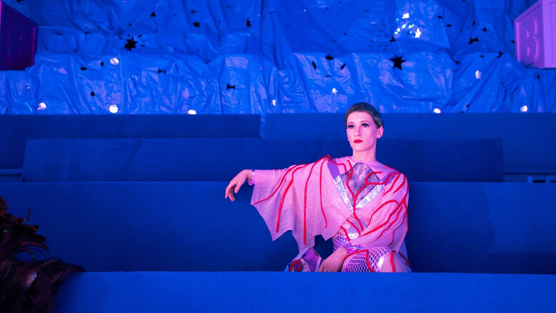 Performer in pink and red costume with blue background for Justene Williams' She Conjured the Clouds