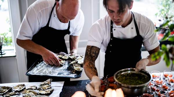 Two men wearing aprons preparing oysters and a sauce. Man in the foreground has tattoos