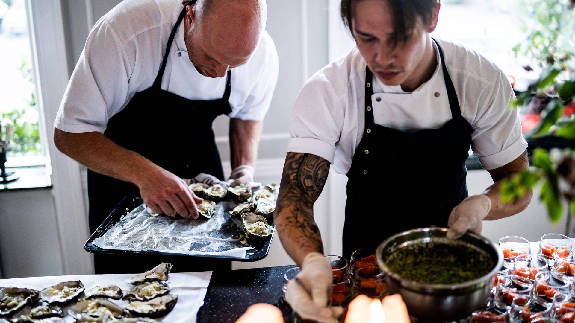 Two men wearing aprons preparing oysters and a sauce. Man in the foreground has tattoos