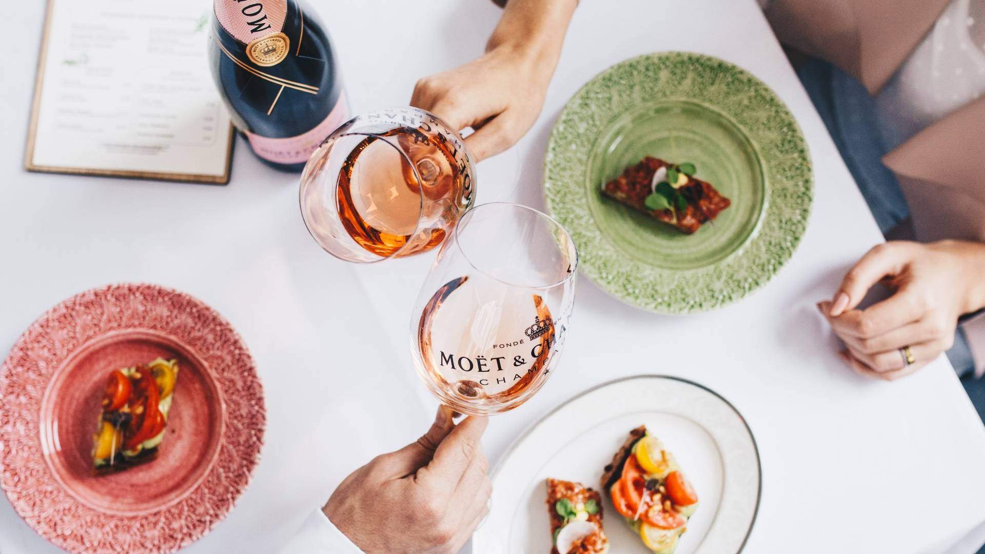 Couple holding glasses in cheers position, glasses branded with Moet & Chandon label with rose champagne