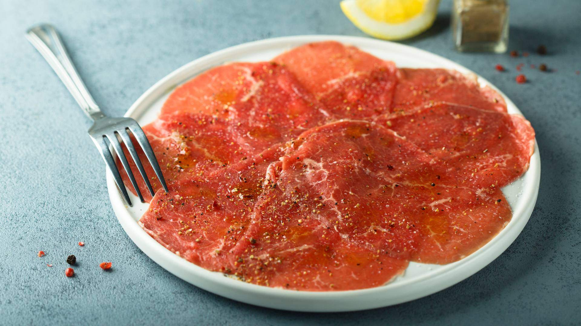 Beef carpaccio on a white plate with fork, lemon and pepper