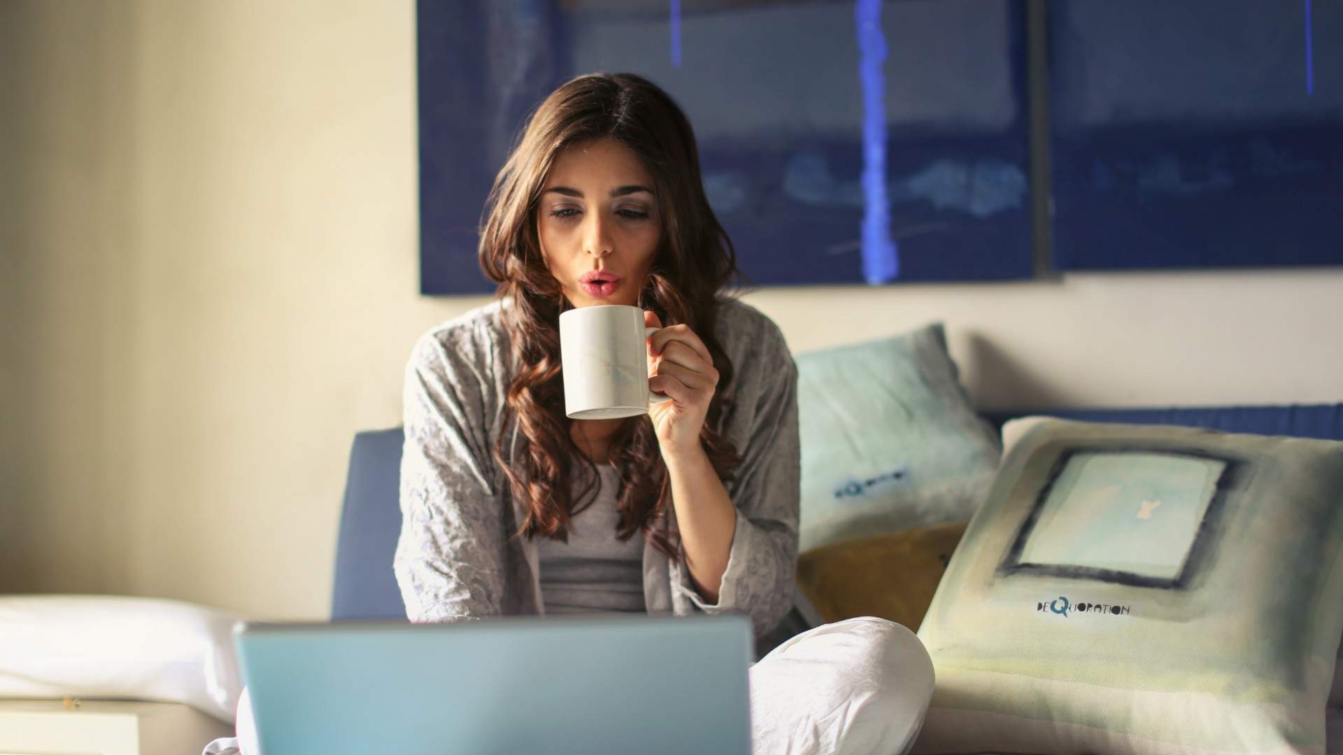 Woman in grey jacket sits on bed drinking coffee