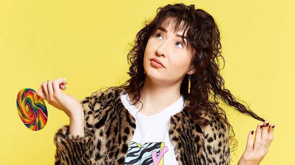 Comedian Bridget Hassed holding a lollipop looking puzzled in front of a yellow backdrop