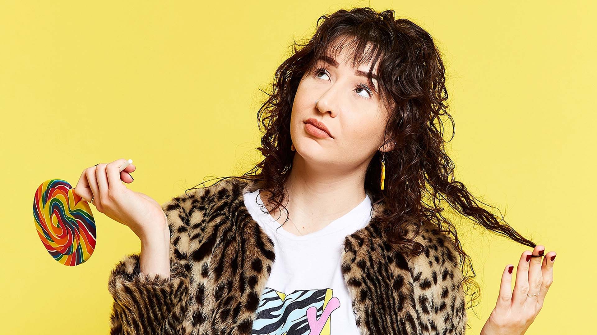 Comedian Bridget Hassed holding a lollipop looking puzzled in front of a yellow backdrop