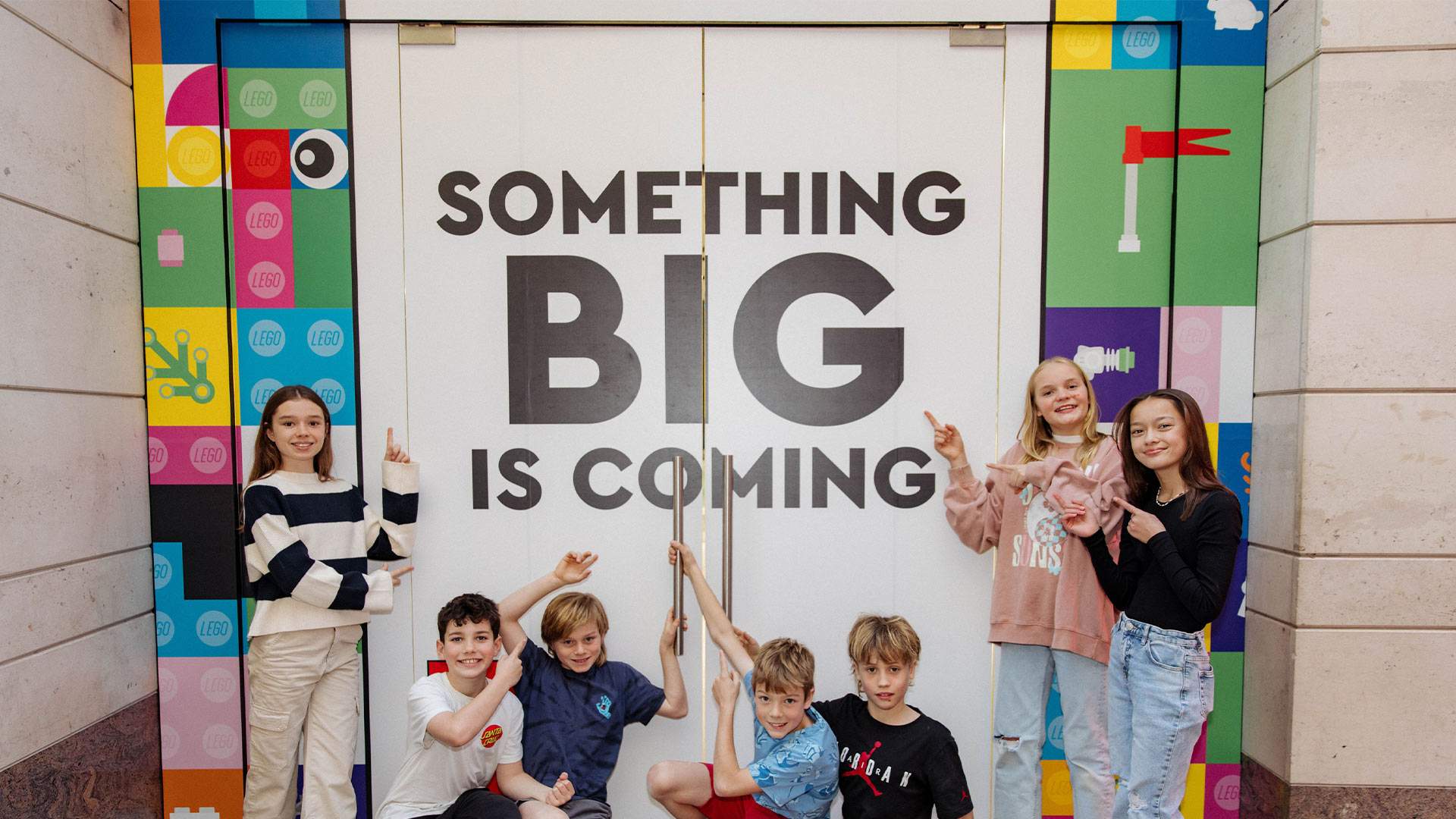 A group of seven kids standing outside Sydney's lego store in front of a sign that says "something big is coming".