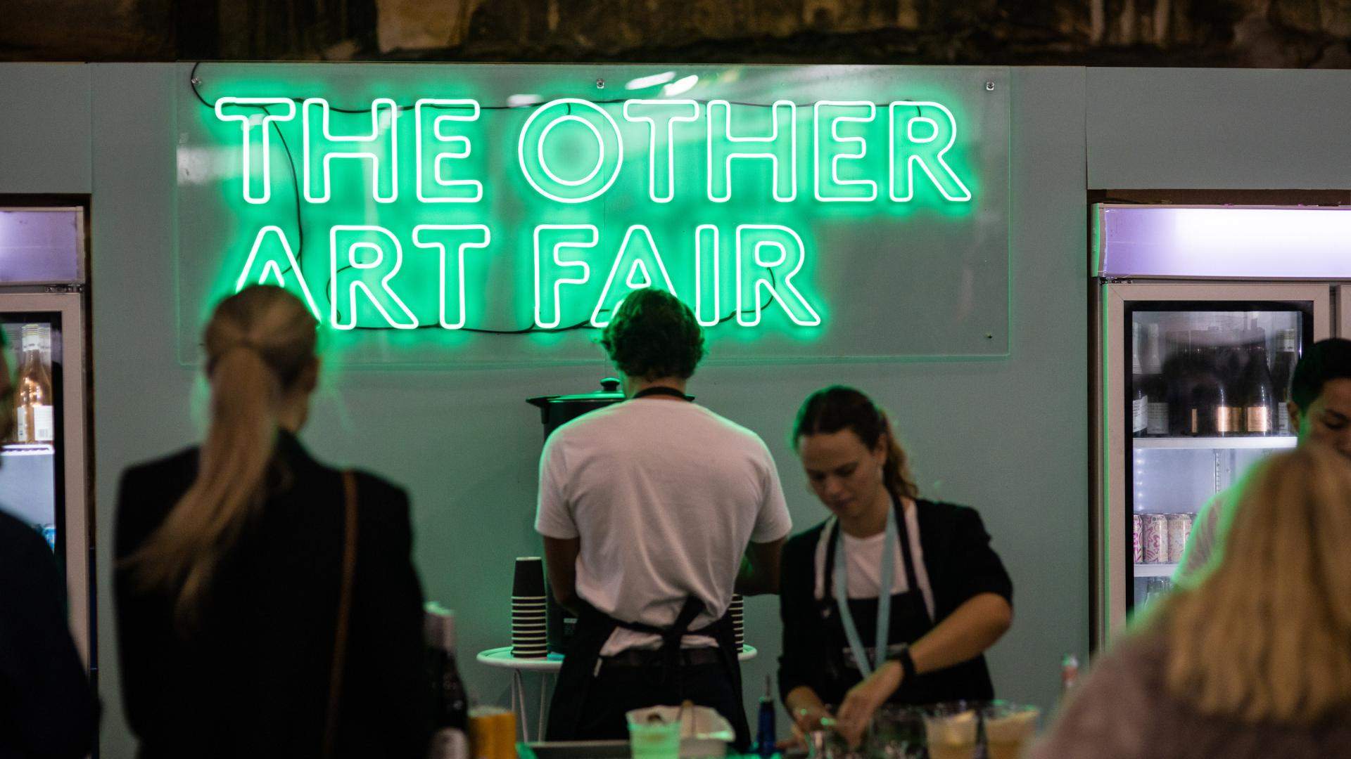 A man is pouring a drink under a sign that says THE OTHER ART FAIR spelled out in bright green LED lights.