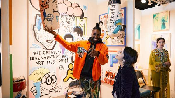 A male artist laughs and points down the barrel of the camera while he holds up a skateboard artwork with a painting of Porky Pig on it.