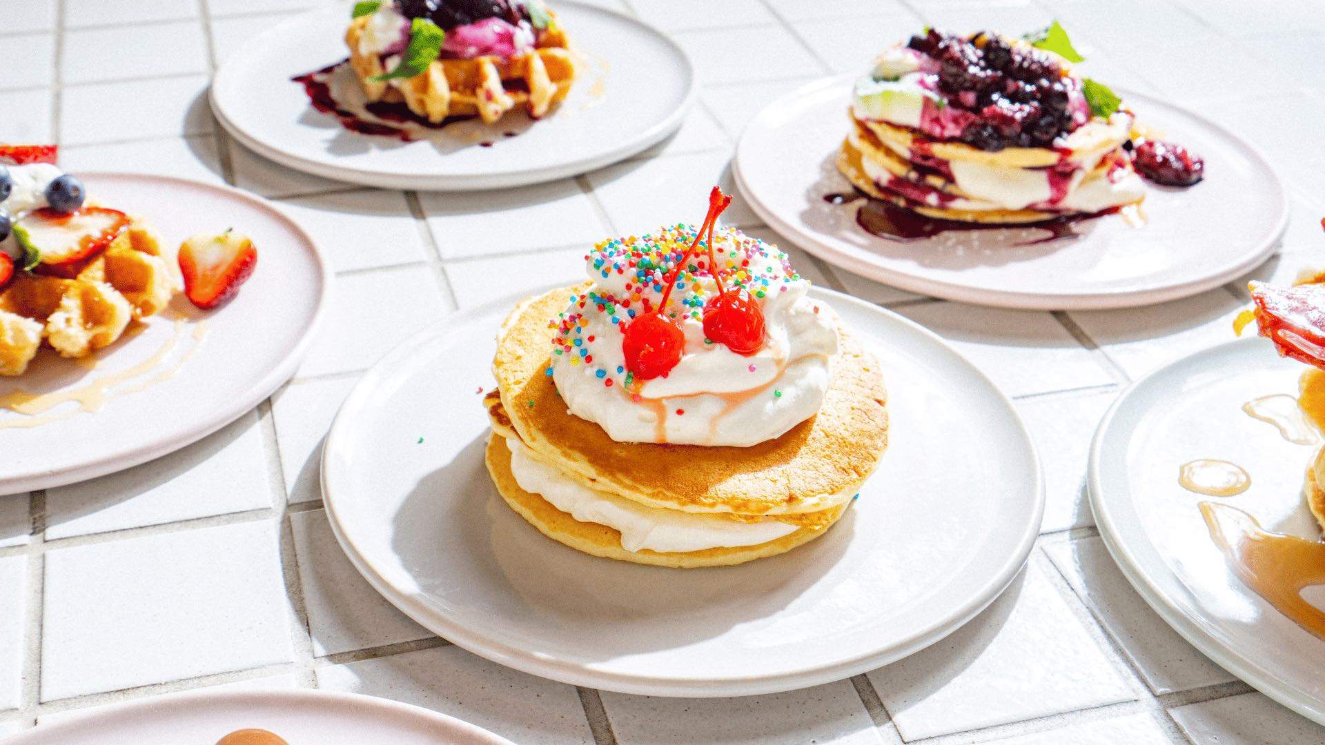 A pancake with whipped cream and cherries surrounded by other pancakes and waffles.