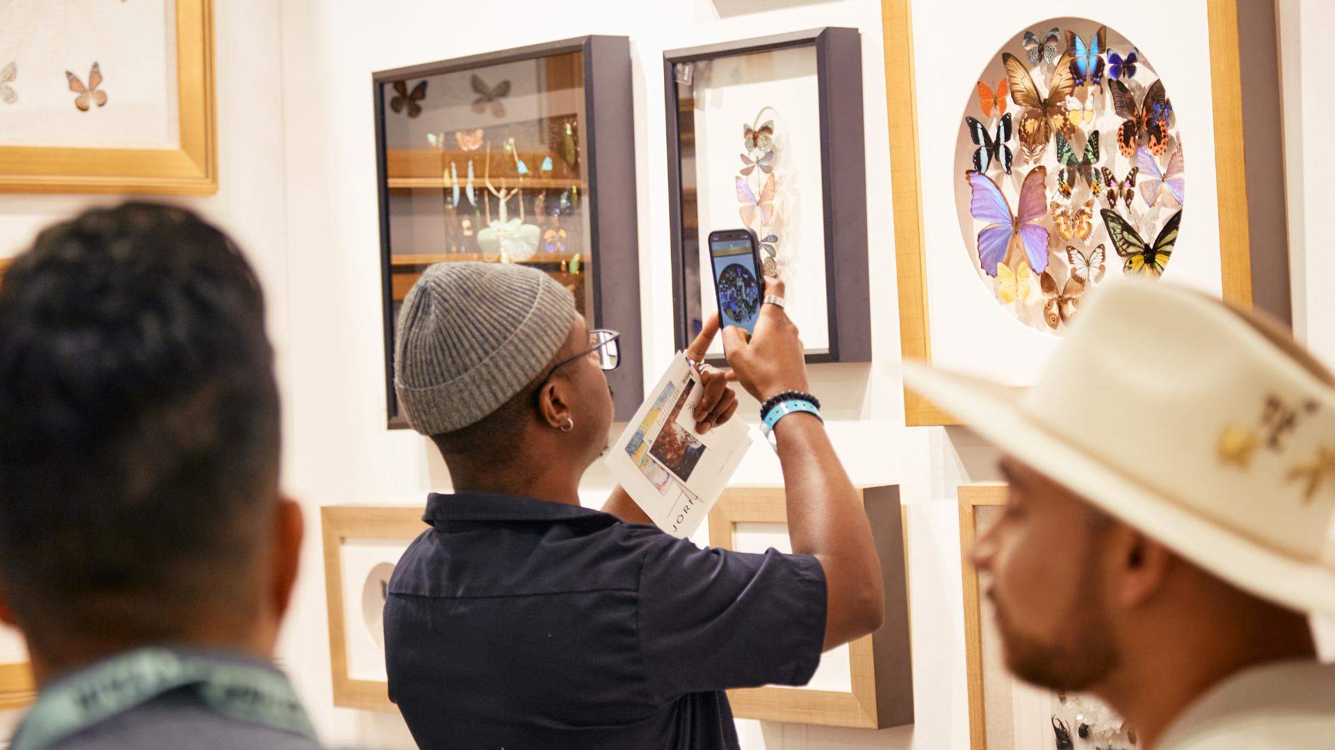 Young man wearing a beanie uses his phone to capture a photo of a framed artwork of multicoloured butterflies at The Other Art Fair.
