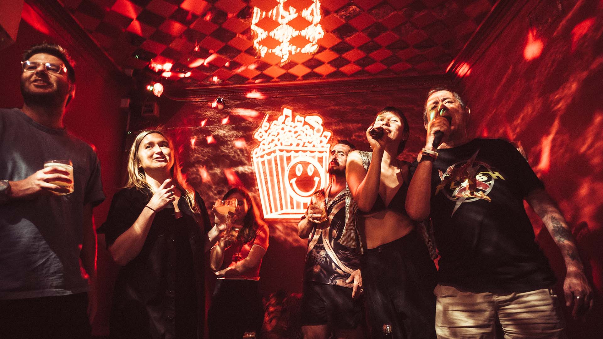 Two young people sing a duet in front of a neon popcorn sign at The Little Guy's red neon-lit karaoke room Poppie's Karaoke