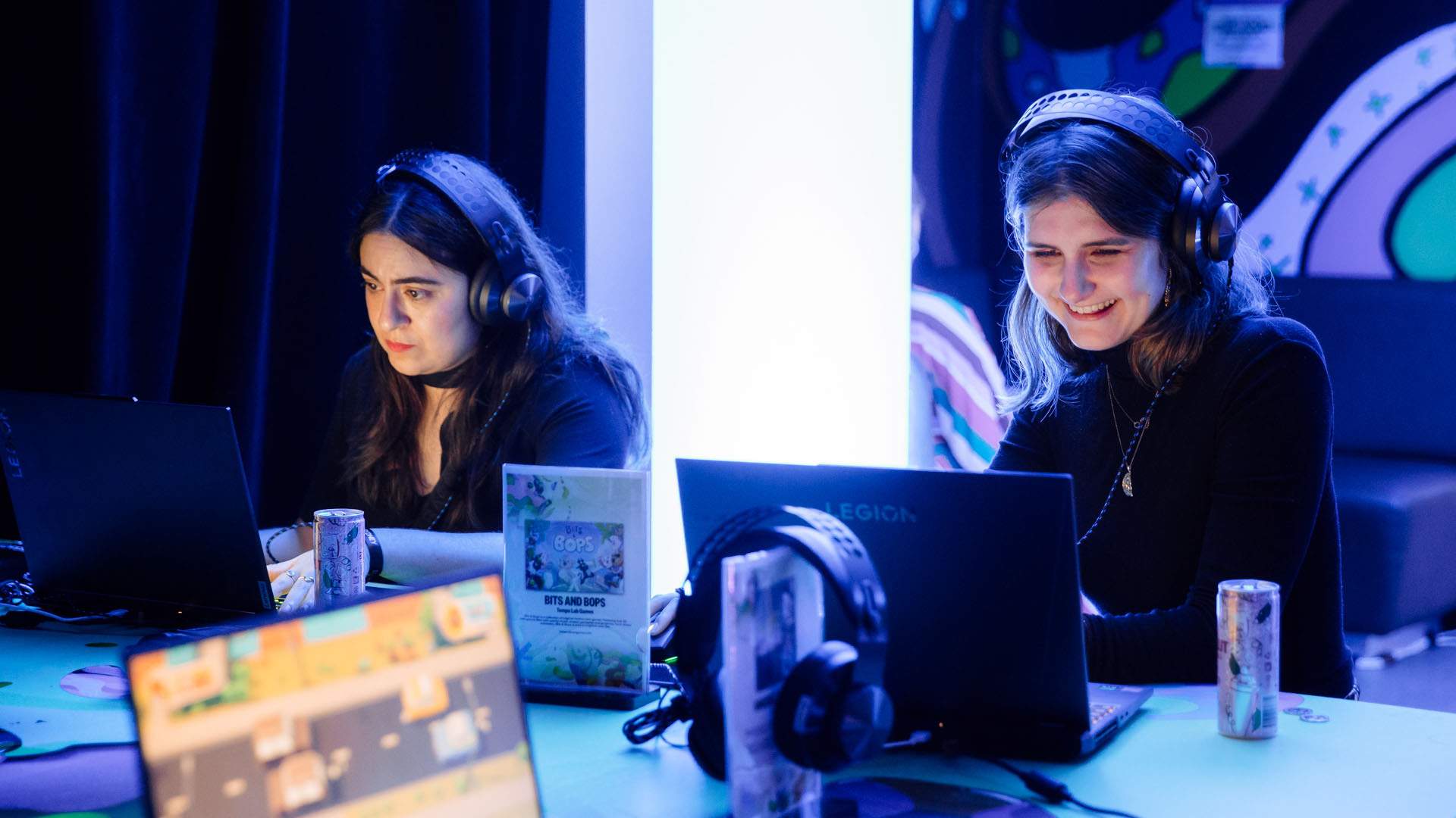 Two women are sitting next to each other with headphones on and gaming on laptops in front of them. They are smiling and obviously having fun.