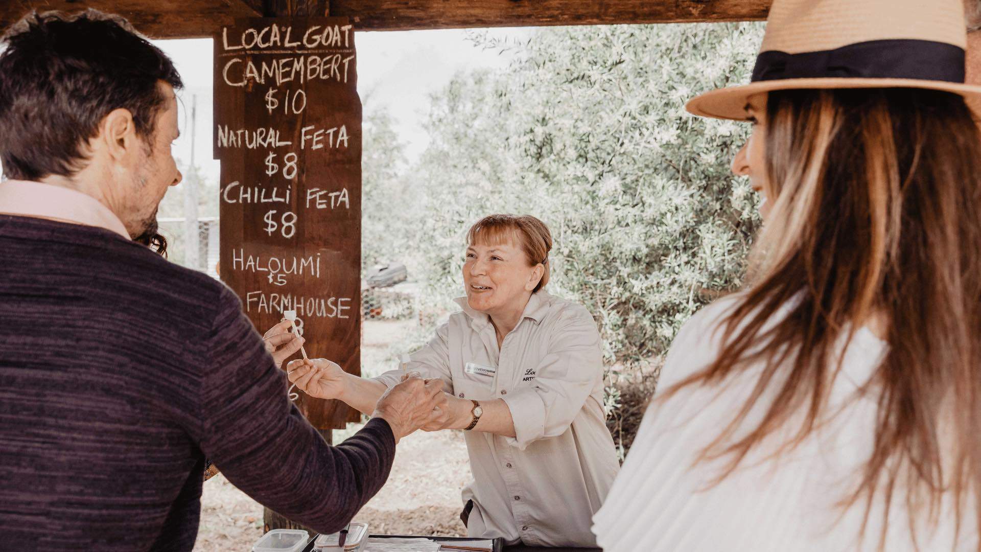 A woman greeting a couple at her cheese stand.