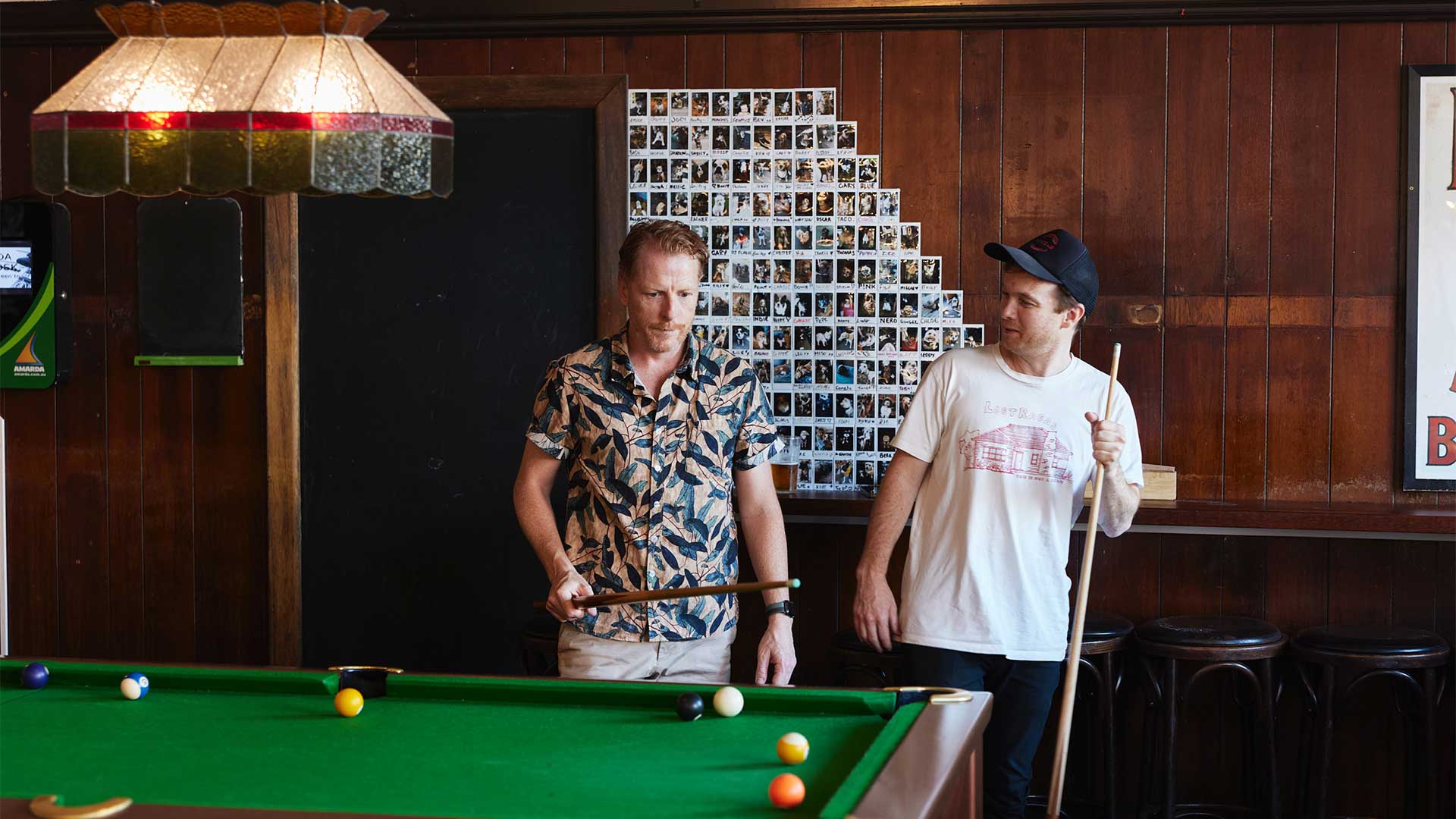 Two men play pool at The Erko in front of a wall of polaroids featuring dogs.