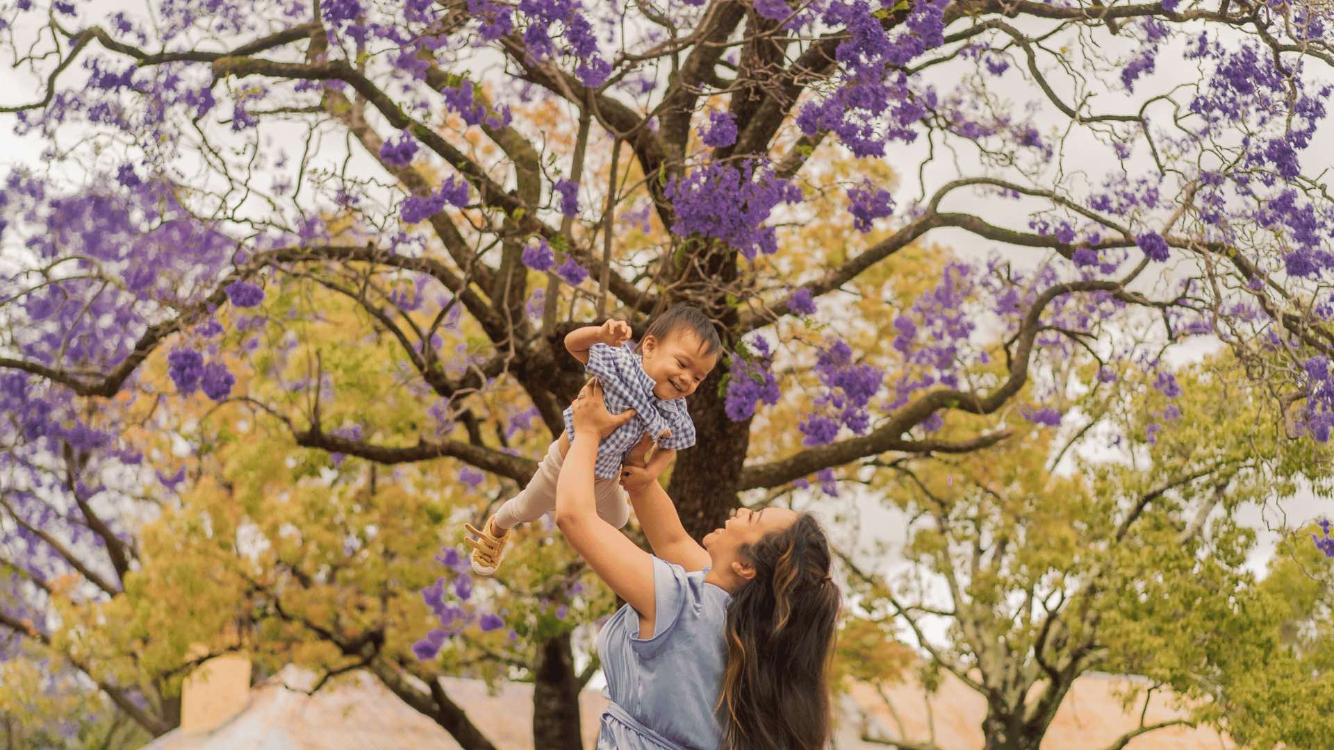 A woman holding her baby in front of a jacaranda tree.
