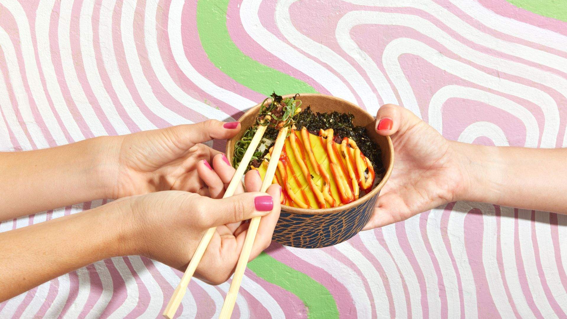 A pair of manicured hands holding a cardboard bowl containing the dish 'The 'Bondi don' which is made of a plant-based patty on a bed of rice, topped with vegan cheese with mustard and tomato sauce squeezed across the top and finished with shredded nori.