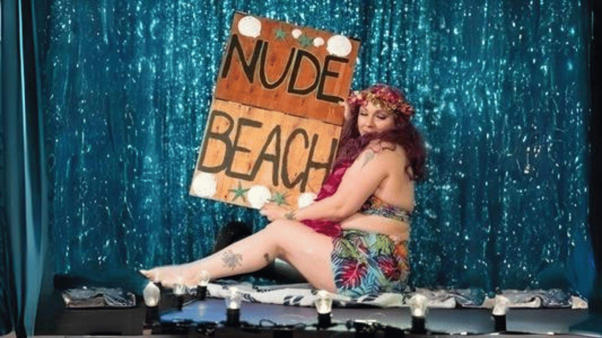 A person holding up a sign reading 'Nude Beach' in front of a sparkly blue backdrop.