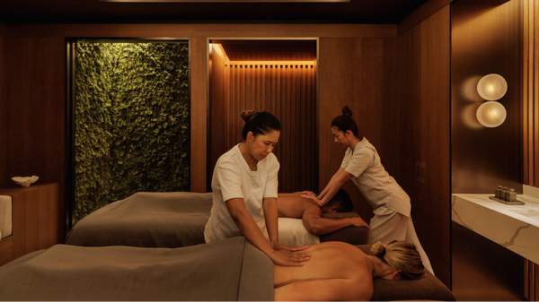 A massage room at The Capella Hotel Sydney's Auriga Spa with two massage therapists giving a massage treatment to a woman lying on a massage table.