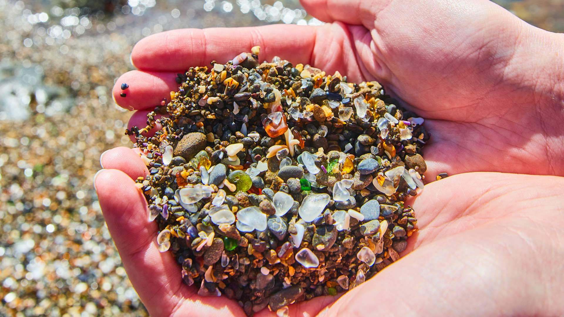 Image of Two hands holding a handful of beach sand filled with shiny and colorful pieces of smooth glass