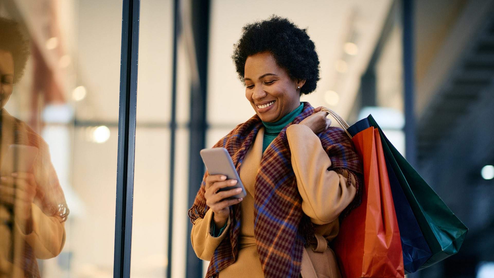 Happy African American woman with shopping bags using mobile phone while standing in front of a store.