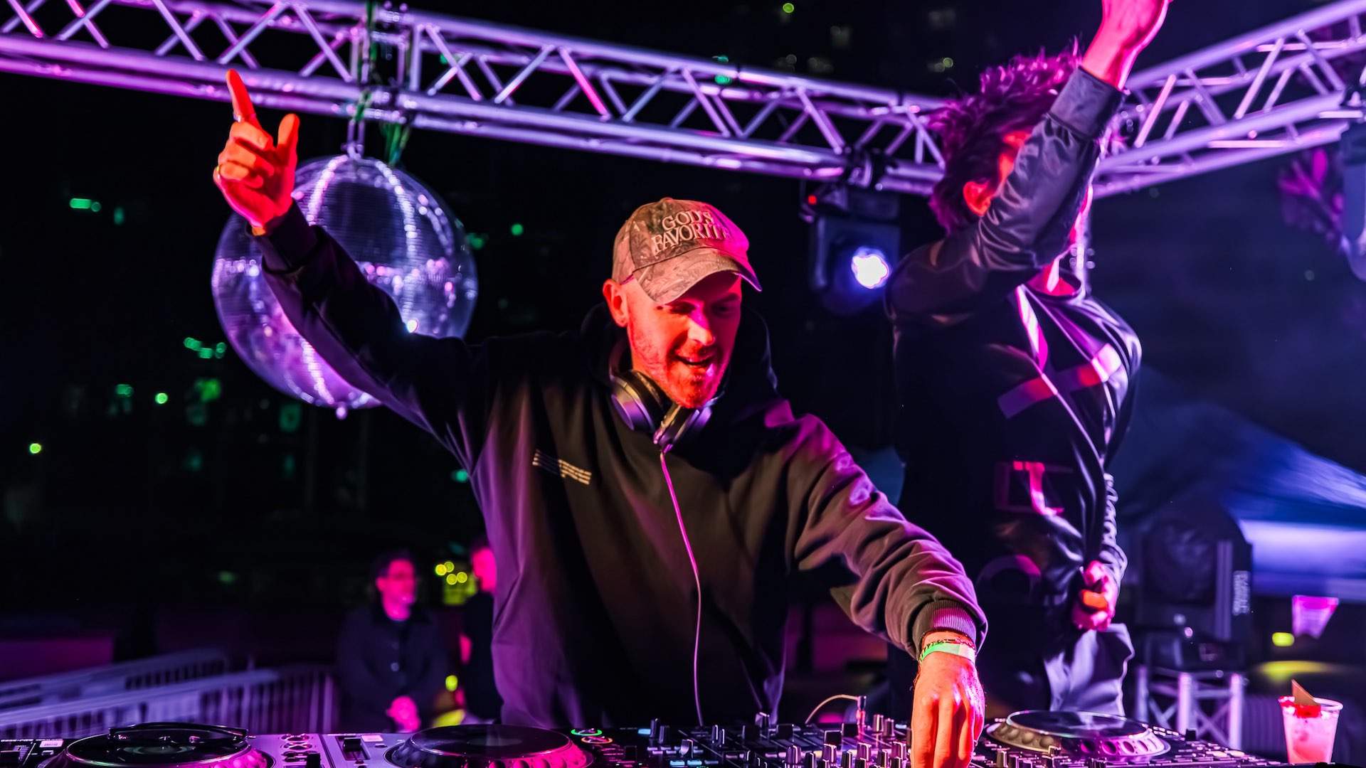 Two DJS, together The Jungle Giants, operate a turntable, each with an arm in the air. A disco ball is illuminated behind them.