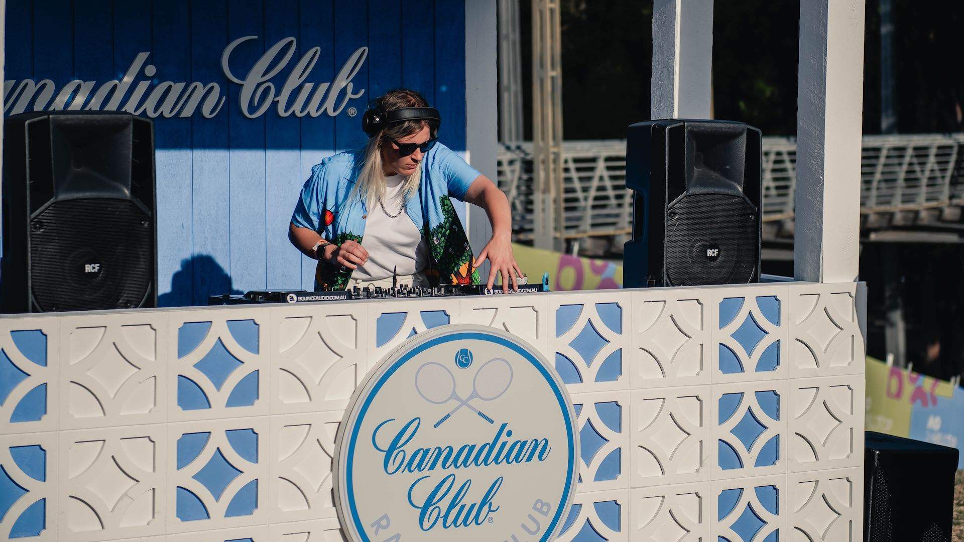 A DJ wearing sunglasses plays behind a blue and white table that says Canadian Club.