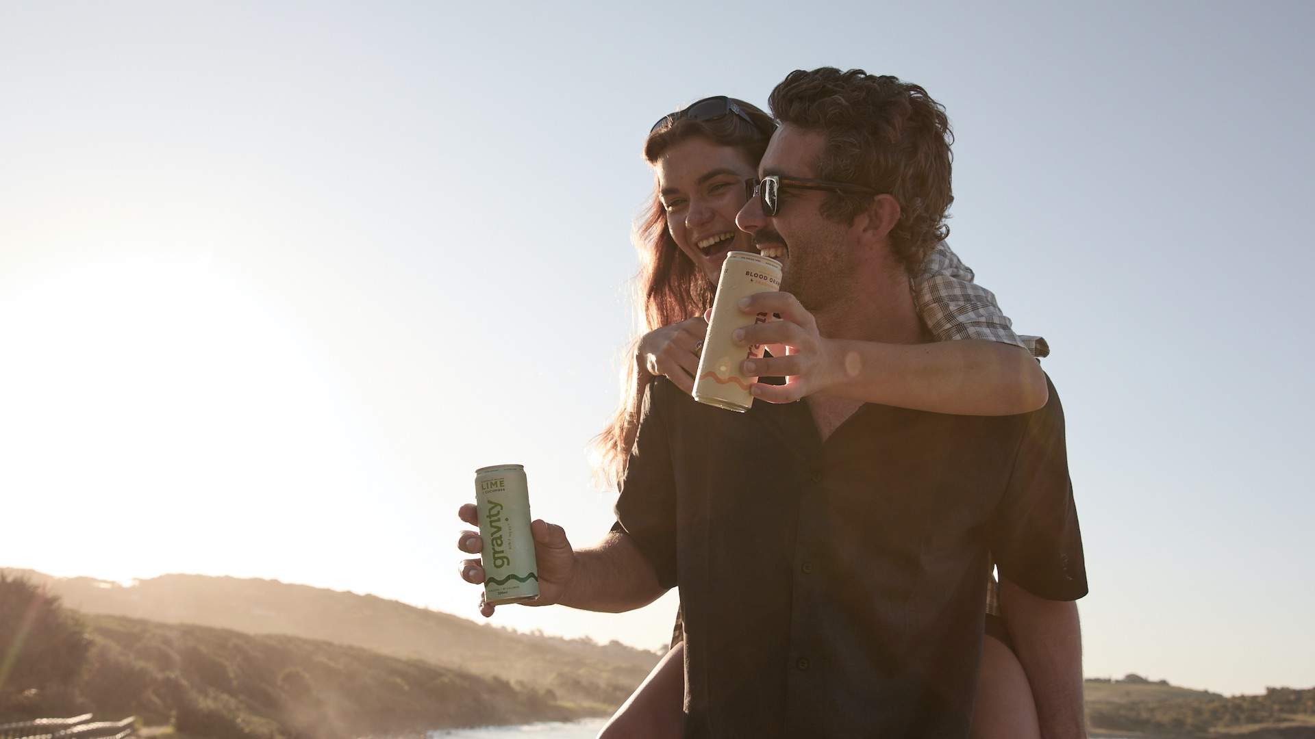 A man piggybacks a woman against a beach landscape, they both hold Gravity Drinks