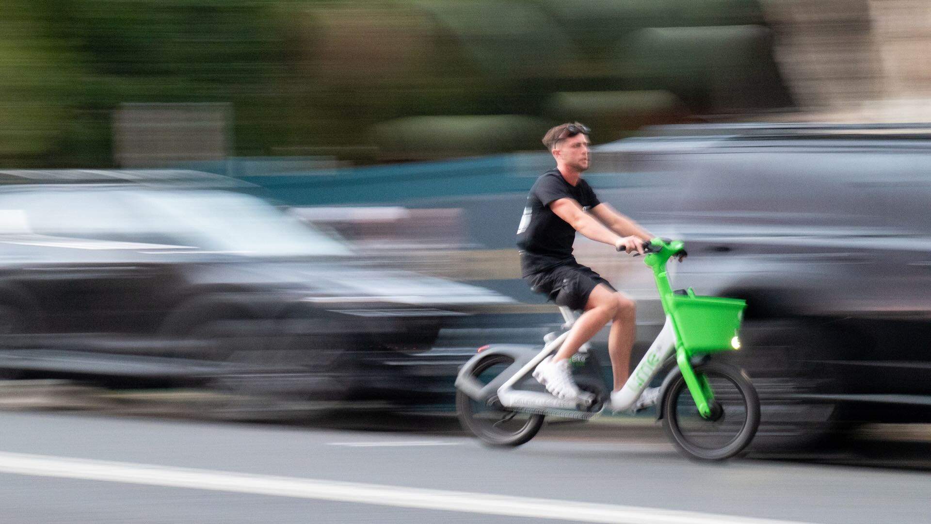 A man riding a share e-bike on Bondi Road, Bondi, heading downhill in the direction of Bondi Beach in the eastern suburbs of Sydney. In the background are parked cars and residential apartment buildings. This image was taken on a hot and sunny evening at sunset on 29 November 2025.