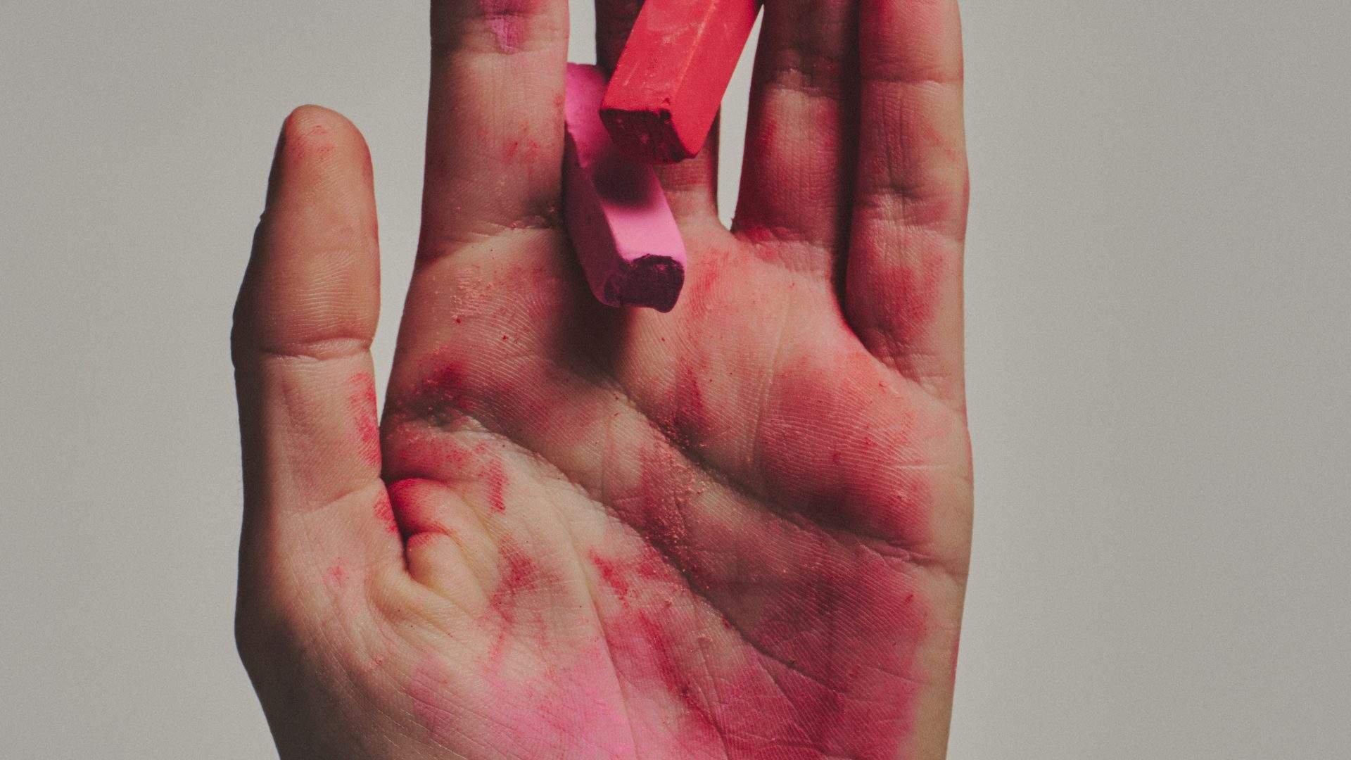 A hand dusted in pink pastel holding two sticks of chalk