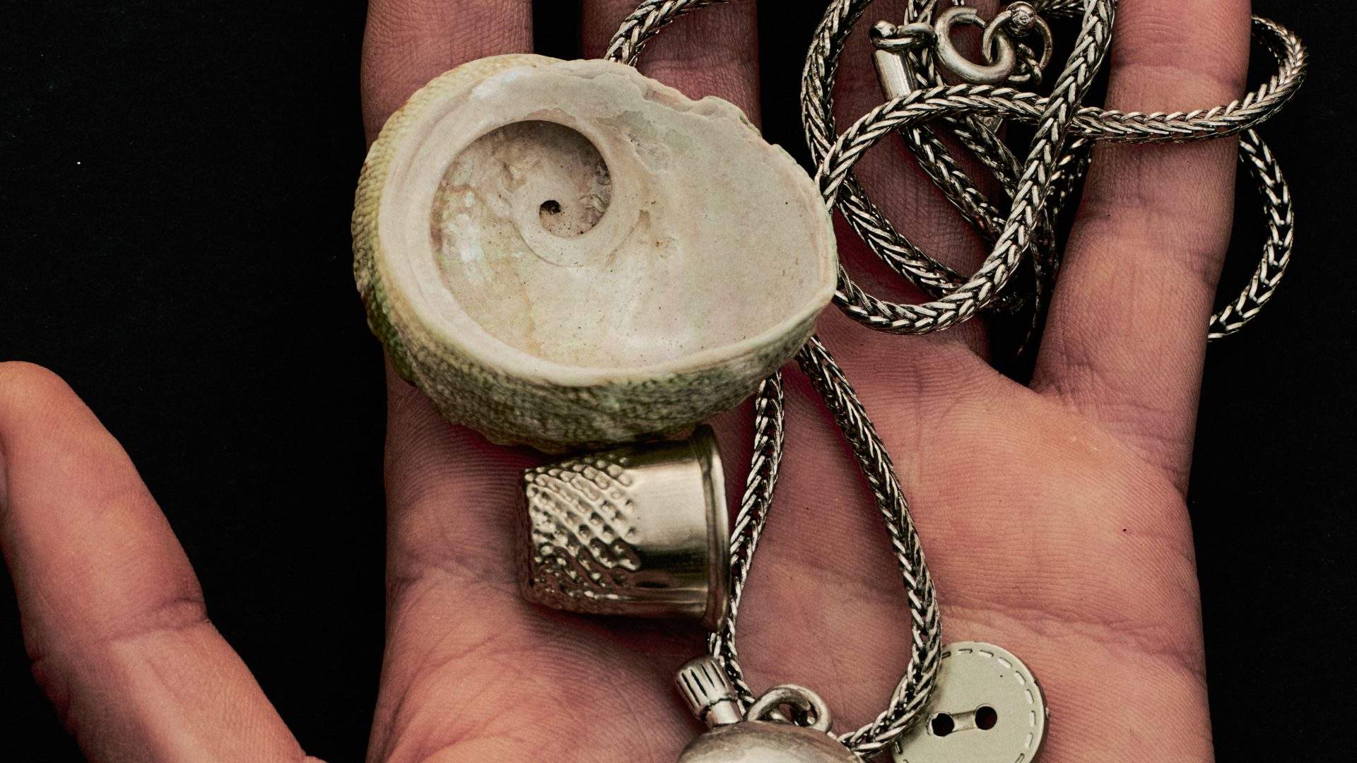 A hand holding a shell, thimble, button and silver chain
