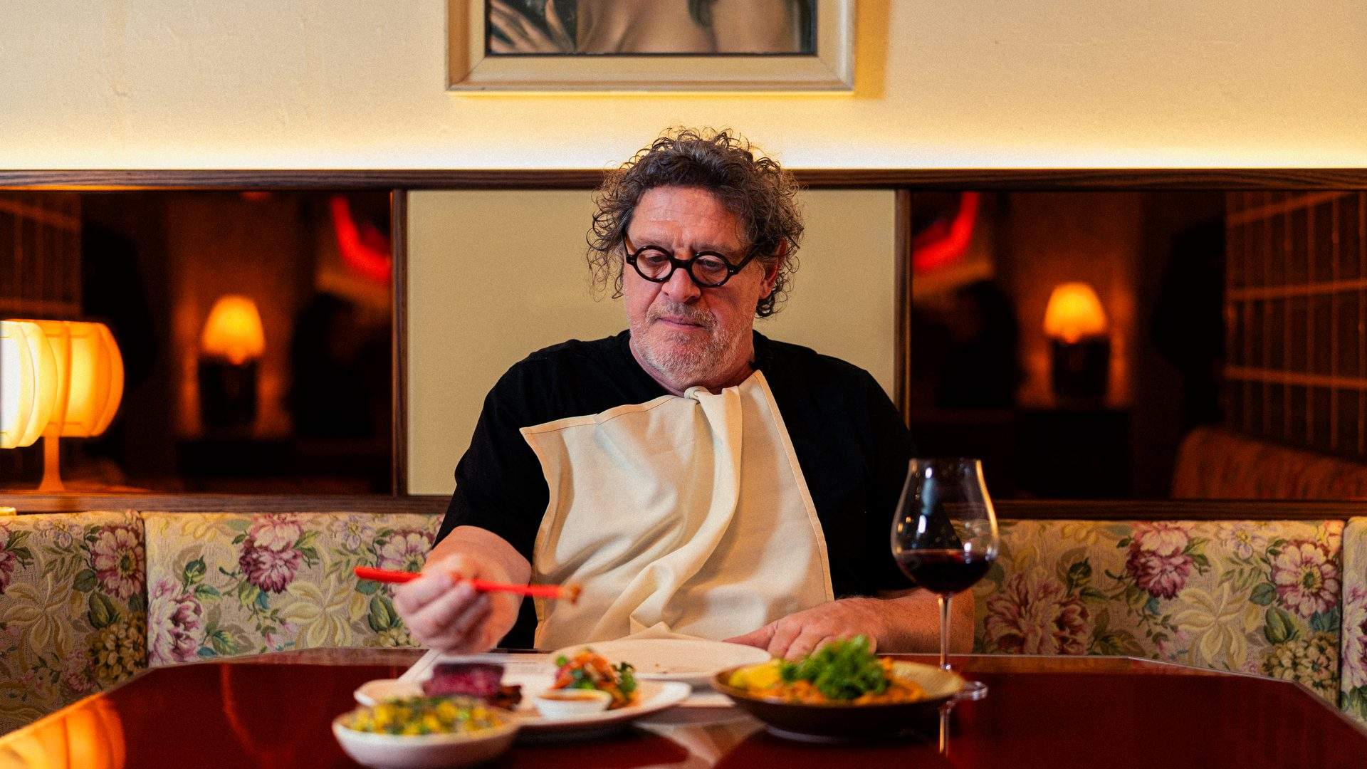 Marco Pierre White seated at a restaurant banquette with a napkin tucked into his collar, a plate of food and a glass of red wine in front of him