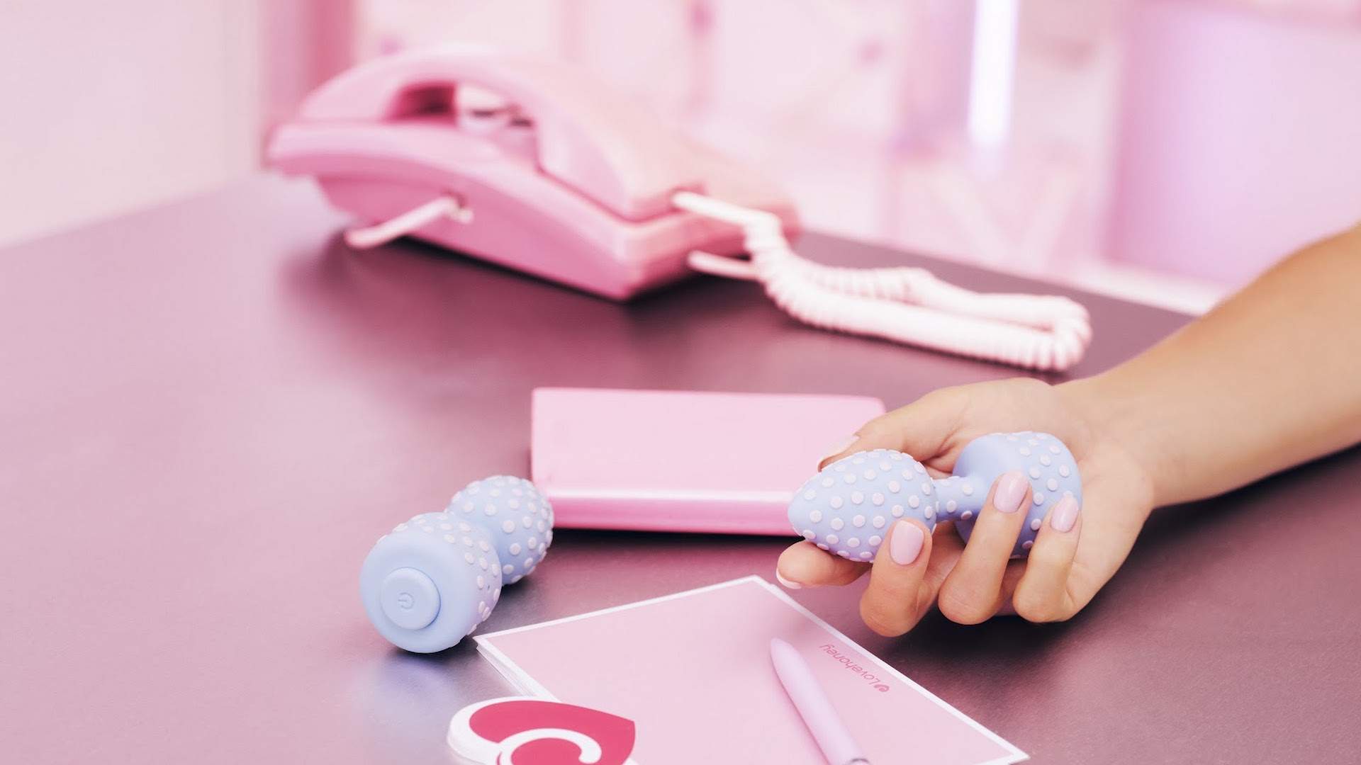 A hand picks up the pale blue polka-dot butt plug from a pink desk