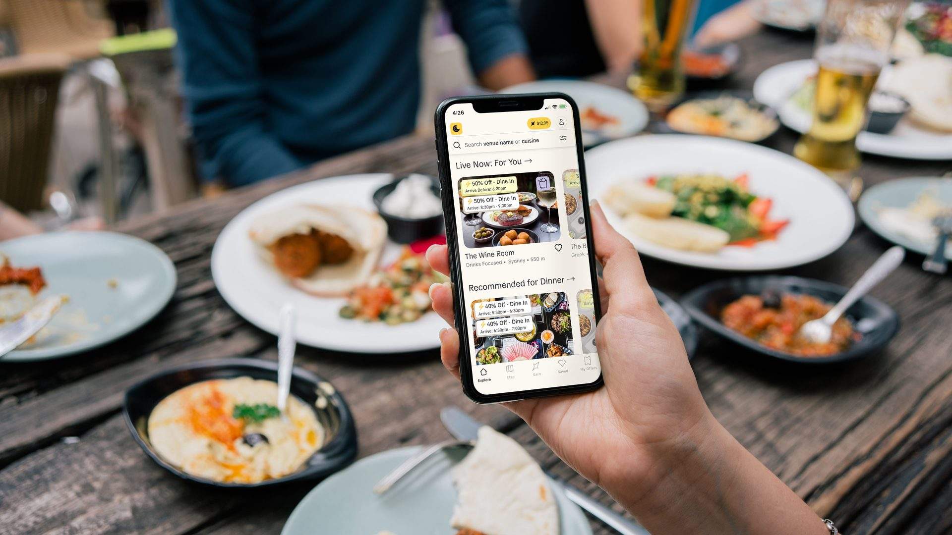 A diner holding a phone showing restaurant deals over a shared table of plates and drinks
